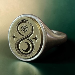 May include: A silver signet ring with an oval face. The face features an engraved design of a snake eating its tail, a sun, a crescent moon, and two stars. The ring has a polished finish.