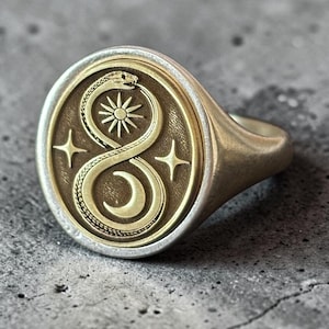 Puede incluir: Un anillo de sello dorado con una cara circular que presenta una serpiente ouroboros grabada formando una figura de infinito. Dentro de la serpiente hay un sol y una luna creciente, flanqueados por dos estrellas de cuatro puntas a los lados.