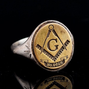 Könnte beinhalten: Ein silberner Ring mit einer goldenen ovalen Oberseite, die ein freimaurerisches Symbol zeigt, einen Zirkel und ein Geodreieck mit einem 'G' in der Mitte und dem Text 'No. 9825'.