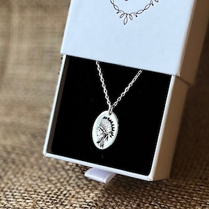 Puede incluir: Un collar de plata con un colgante ovalado que presenta un diseño de tocado nativo americano. El collar está en una caja blanca con un interior negro.