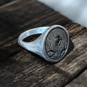 May include: A silver signet ring with a detailed bird and branch design. The ring has a brushed metal finish and a round face with an engraved bird motif. The ring is displayed on a wooden surface.