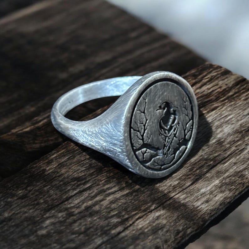 May include: A silver signet ring with a detailed bird and branch design. The ring has a brushed metal finish and a round face with an engraved bird motif. The ring is displayed on a wooden surface.