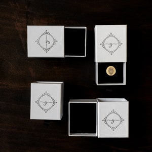 May include: White jewelry boxes with black interiors are arranged on a dark wood surface. Each box features a black and white floral design with a letter. One box contains a gold-colored round pin with a crown and the letter "M".