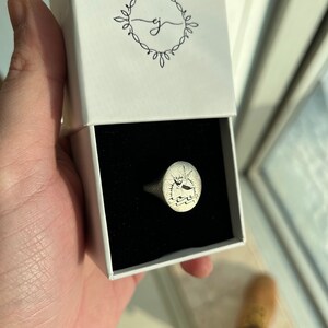 May include: A silver signet ring with a detailed design of a ship sailing on the water. The ring is in a white box with a black interior.