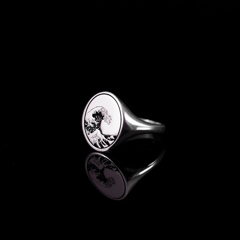 May include: A silver signet ring with a black and white image of a wave on the face of the ring.
