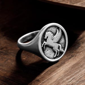 May include: A silver signet ring featuring a detailed relief of a Pegasus, a mythical winged horse. The ring has a rounded face with the Pegasus design and a smooth, polished band. The ring is set against a dark wood surface.