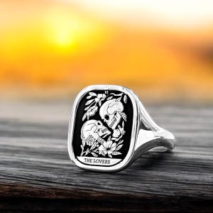 K&ouml;nnte beinhalten: Ein silberner Ring mit einem schwarzen quadratischen Einsatz, der ein Tarotkarten-Design mit zwei sich umarmenden Skeletten mit Blumen zeigt. Die Karte lautet "THE LOVERS".