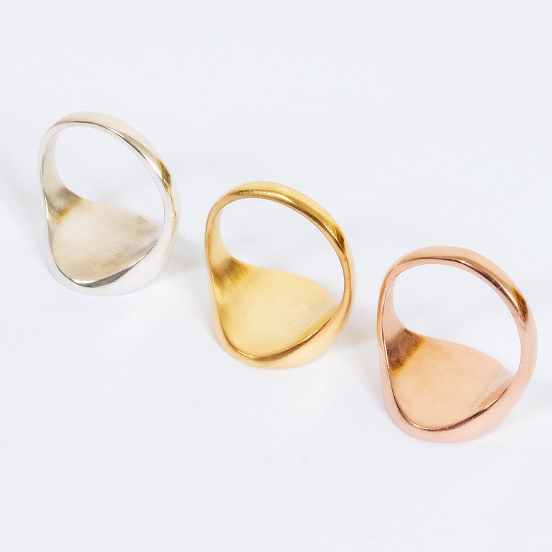 May include: Three signet rings in silver, gold, and rose gold. Each ring has a smooth, oval face and a rounded band. The rings are displayed on a white surface, showcasing their different metallic finishes.