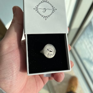 May include: A silver signet ring with an oval face featuring a detailed engraved design. The ring is displayed in a white box with a black interior.