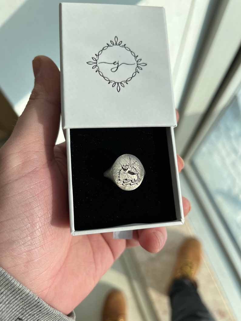 May include: A silver signet ring with an oval face featuring a detailed engraved design. The ring is displayed in a white box with a black interior.