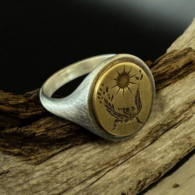 May include: A silver ring with a brushed finish and a circular, brass-colored face. The face features a sun design with rays, a bird, and a wheat-like pattern. The ring is set against a wooden background.