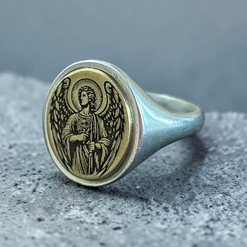 May include: Silver signet ring with a gold-coloured oval face. The face features a detailed engraving of an angel with wings, holding a key. The ring has a polished finish and a classic design.