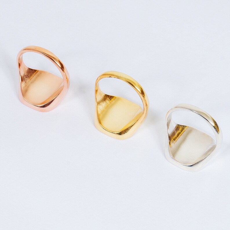 May include: Three signet rings in different metal finishes: rose gold, gold, and silver. The rings are all the same design, with a square-shaped top and a rounded band.