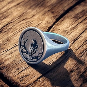 May include: Silver signet ring featuring a detailed raven design within a circular frame. The raven is perched on a branch, set against a textured background. The ring has a polished finish and a sturdy band, perfect for everyday wear.