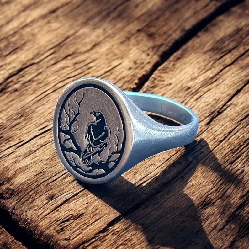 May include: Silver signet ring featuring a detailed raven design within a circular frame. The raven is perched on a branch, set against a textured background. The ring has a polished finish and a sturdy band, perfect for everyday wear.