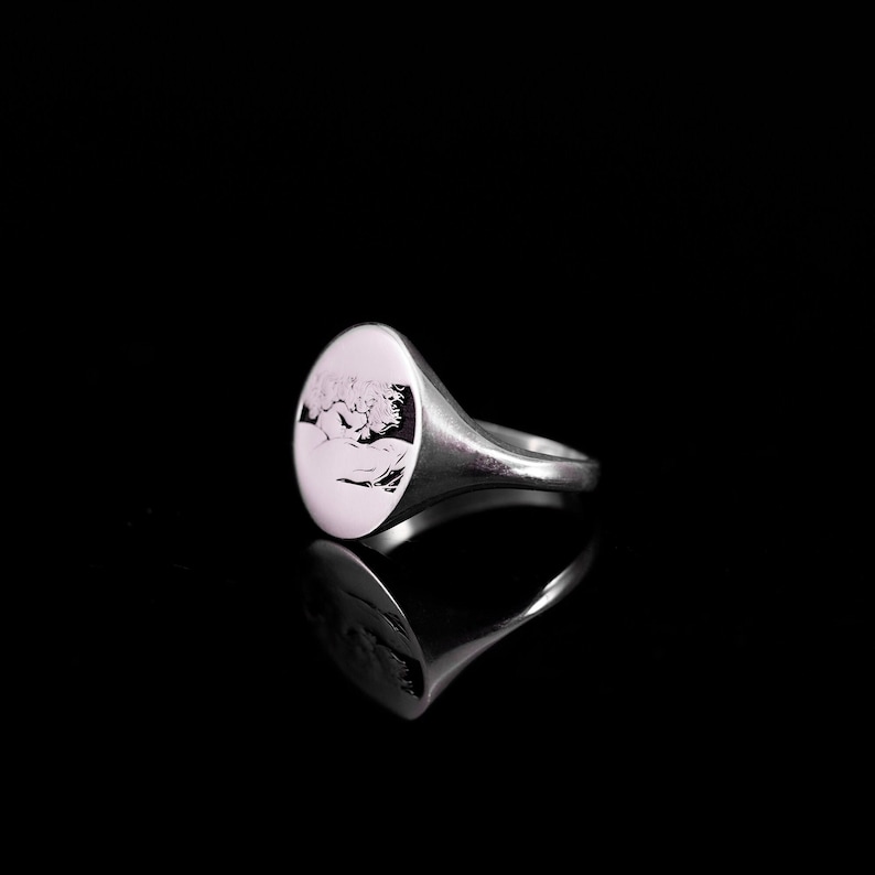 May include: A silver signet ring with a round, engraved top featuring a detailed design. The ring has a smooth, polished finish.
