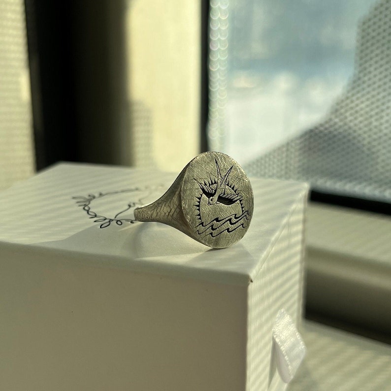 May include: A silver signet ring with a bird flying over waves engraved on the top. The ring is sitting on a white box.