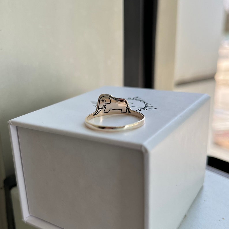 Puede incluir: Un anillo de oro con un dise&ntilde;o de elefante en la parte superior. El anillo est&aacute; sobre una caja blanca con la palabra "absolutamente" escrita en ella.