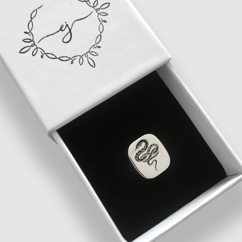 May include: A silver ring with a black snake design in a white box with a black interior.