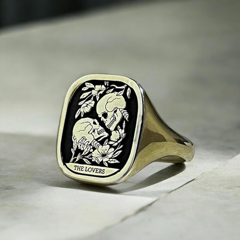 K&ouml;nnte beinhalten: Ein goldener Siegelring mit einer schwarzen quadratischen Fl&auml;che, die eine wei&szlig;e Illustration von zwei sich umarmenden Skeletten zeigt. Der Text "THE LOVERS" und die r&ouml;mische Zahl "VI" sind auf der Ringfl&auml;che eingraviert.