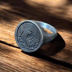 May include: A silver signet ring with an oval face featuring a detailed relief of a bird perched in a natural setting. The ring has a smooth, polished band and a dark, antiqued finish, set against a wooden surface.