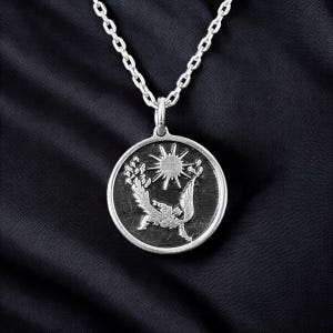 May include: Silver-toned pendant necklace featuring a circular pendant with a detailed relief design. The pendant depicts a sun, bird, and floral elements. The necklace has a delicate chain and is set against a dark background.