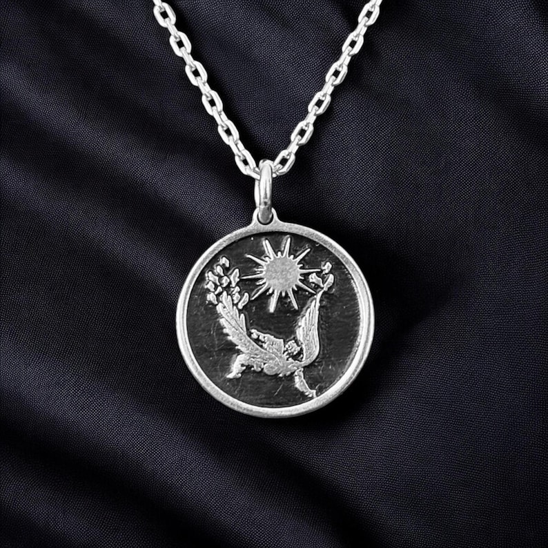 May include: Silver-toned pendant necklace featuring a circular pendant with a detailed relief design. The pendant depicts a sun, bird, and floral elements. The necklace has a delicate chain and is set against a dark background.