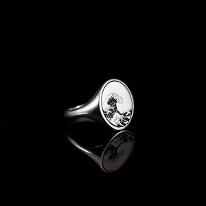 May include: A silver signet ring with a detailed engraving of a wave. The ring is oval shaped and has a smooth, polished finish.