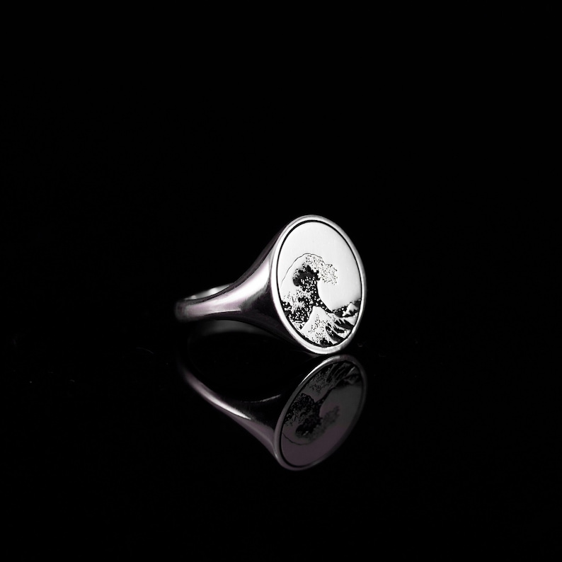 May include: A silver signet ring with a detailed engraving of a wave. The ring is oval shaped and has a smooth, polished finish.