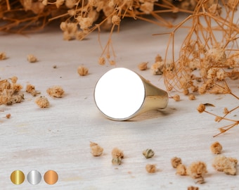 Könnte beinhalten: Ein goldener Siegelring mit einer leeren ovalen Oberseite. Der Ring liegt auf einer weißen Oberfläche mit verstreuten getrockneten Blumen.