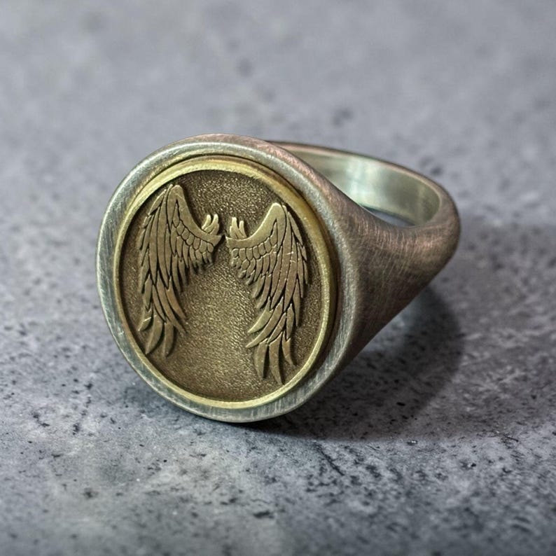 May include: A silver ring with a brushed finish. The ring features a circular, bronze-colored center with an embossed design of two symmetrical wings. The ring is set against a gray background.