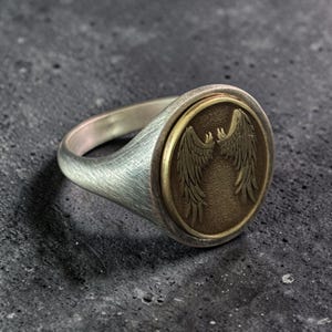 May include: A silver ring with a brushed finish and a circular, bronze-colored emblem. The emblem features a detailed design of stylized wings. The ring is set against a dark, textured background.