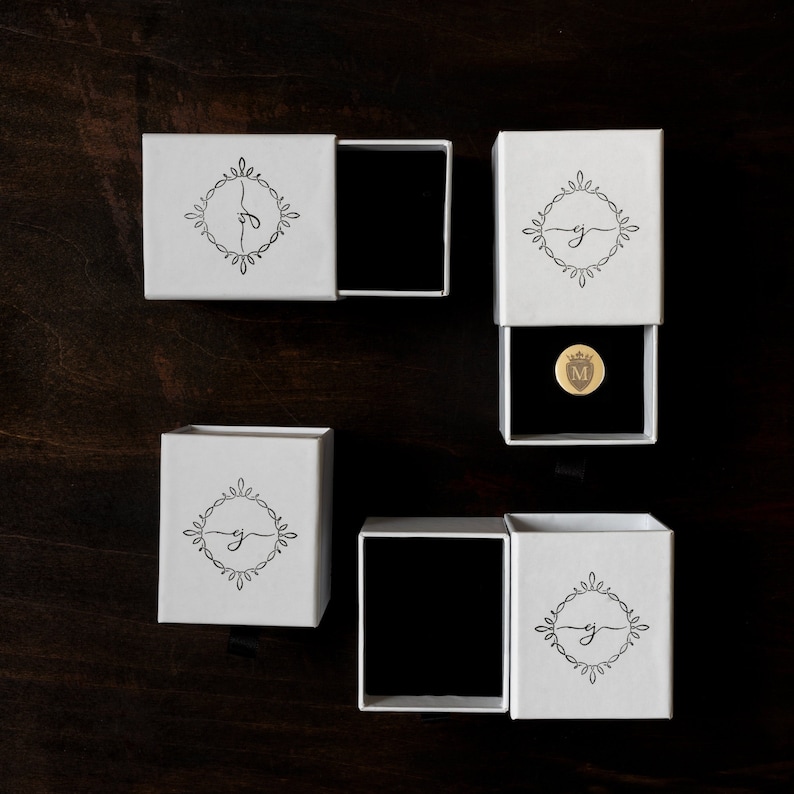 Pu&ograve; includere: Quattro scatole bianche con bordi neri e un design monogramma in bianco e nero. Le scatole sono aperte per rivelare un interno in velluto nero e una moneta d'oro con la lettera "M" su di essa.