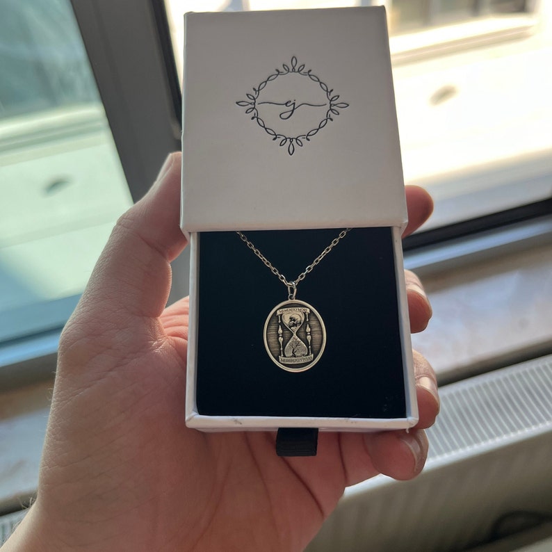 Pu&ograve; includere: Una collana d'argento con un ciondolo ovale che presenta un design a clessidra e l'iscrizione "Il tempo non aspetta nessuno". La collana &egrave; in una scatola bianca con un interno nero.