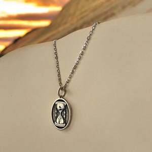 Pu&ograve; includere: Una collana con catena d'argento con un ciondolo rotondo che presenta un design a clessidra in bianco e nero con il testo "Memento Mori" in alto e in basso.