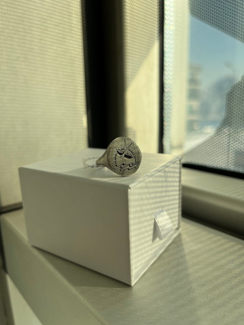 May include: A silver signet ring with an engraved design. The ring is in a white box with a white ribbon.