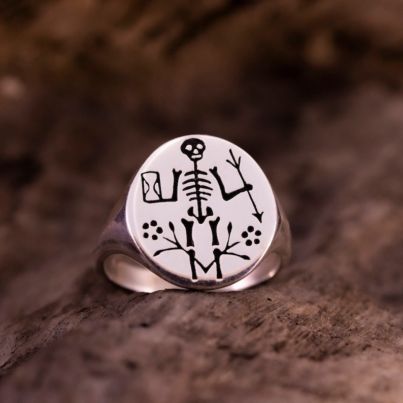 May include: A silver signet ring with a black and white engraved image of a skeleton holding a spear and a small object. The ring is on a brown wooden surface.