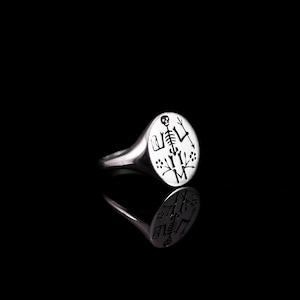 May include: A silver signet ring with a round, engraved top featuring a skeleton with a skull, surrounded by small floral designs.