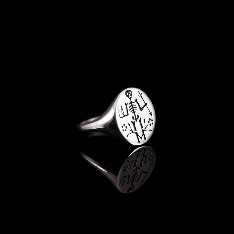 May include: A silver signet ring with a round, engraved top featuring a skeleton with a skull, surrounded by small floral designs.