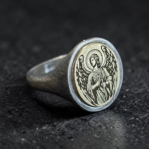 May include: Silver ring with an engraved angel design. The ring has a round, gold-coloured centre with an angel figure, surrounded by a silver band. The angel has wings and is holding an object. The ring is set against a dark background.