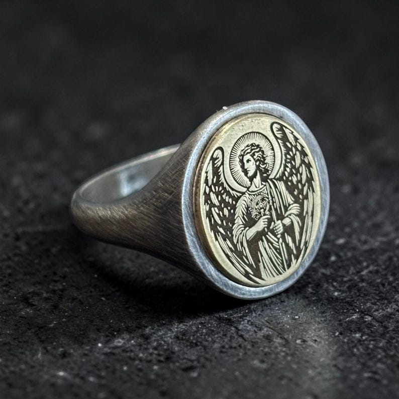 May include: Silver ring with an engraved angel design. The ring has a round, gold-coloured centre with an angel figure, surrounded by a silver band. The angel has wings and is holding an object. The ring is set against a dark background.
