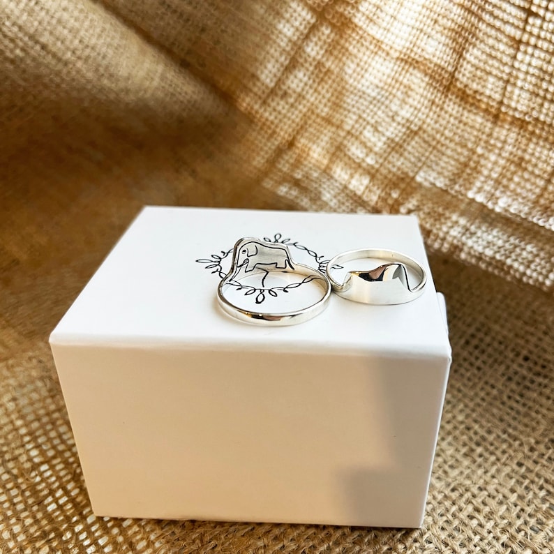 Puede incluir: Tres anillos de plata en una caja blanca. Los anillos son de dise&ntilde;o simple y moderno. Un anillo presenta una silueta de elefante.