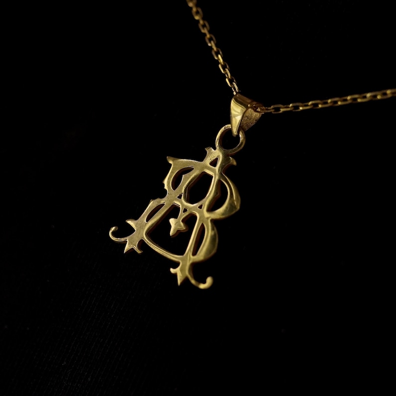 May include: A gold pendant necklace with a detailed, ornate design. The pendant features a stylized letter 'B' with intricate flourishes.