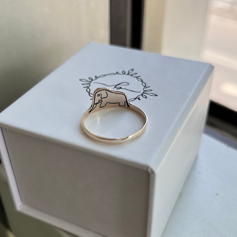 Puede incluir: Un anillo de oro con un dise&ntilde;o de elefante. El anillo est&aacute; en una caja blanca con la palabra "belle" escrita en cursiva.