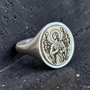 May include: A silver ring with a circular face depicting an engraved angel with wings and a halo. The angel is in a detailed, vintage style. The ring has a smooth, polished finish.