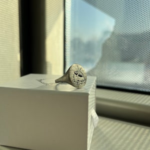 May include: A silver signet ring with a design of a boat on a wave. The ring is sitting on a white box.