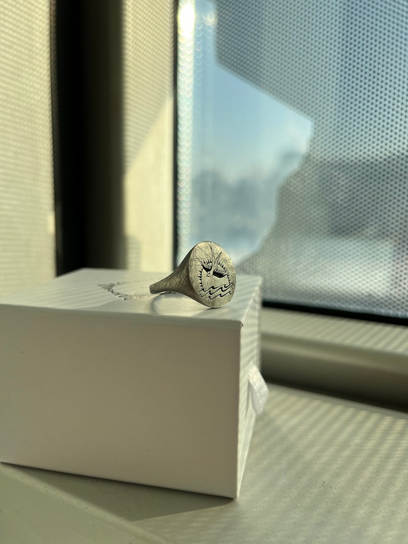 May include: A silver signet ring with a design of a boat on a wave. The ring is sitting on a white box.
