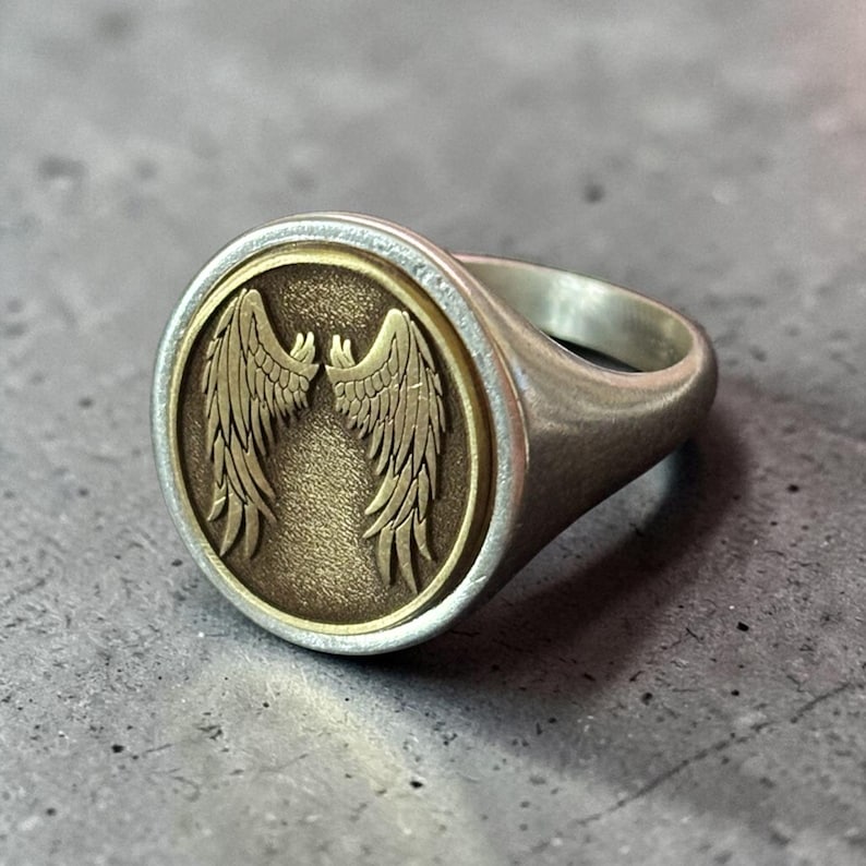 May include: Silver ring with a circular face featuring a detailed design of two symmetrical wings in a bronze color. The ring has a polished finish and a classic signet style.