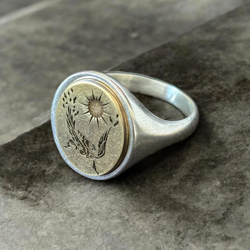 May include: Silver signet ring with an oval face. The face features a gold-colored center with an engraved design of a sun and stylized plant. The ring has a polished finish.