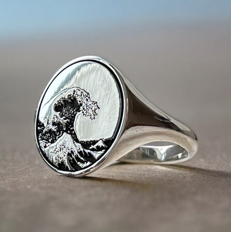 May include: A silver ring with an oval-shaped face featuring an engraved image of the Great Wave off Kanagawa, a famous Japanese woodblock print by Hokusai.